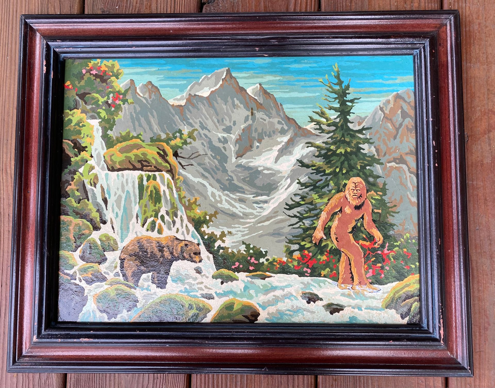 A nature painting with mountains in the background, a bear crossing a river and Big Foot staring at the bear. 