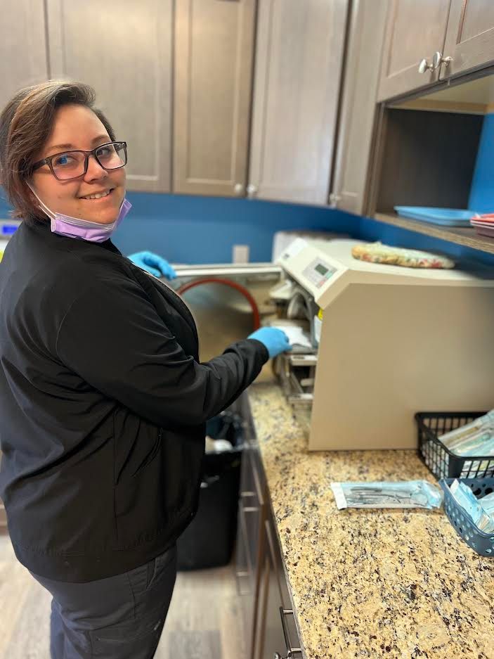 Woman in scrubs at a dental office, using a sterilizer, smiling. Blue gloves, gray cabinets, and countertops.