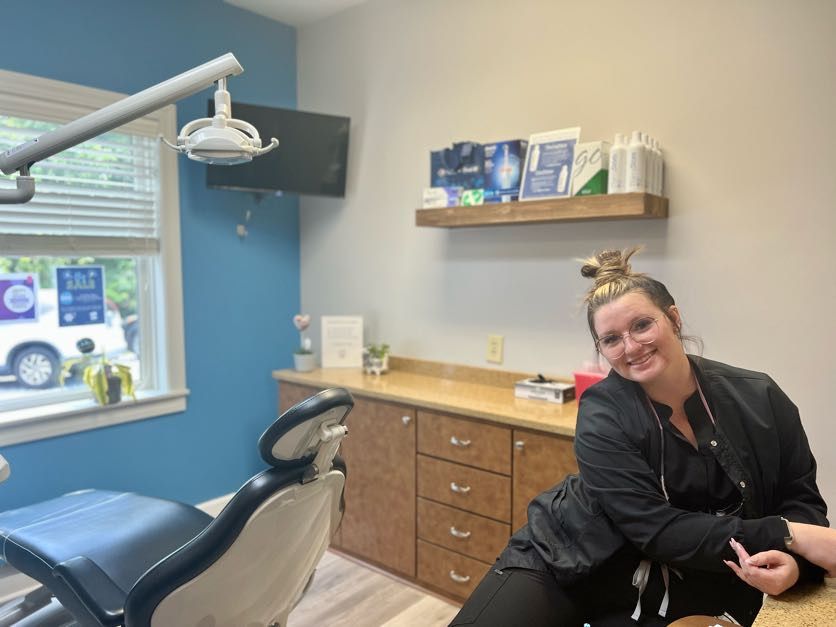Dental office room; smiling woman in scrubs leans on countertop, blue wall, equipment.