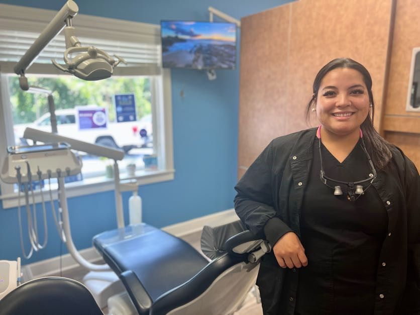 A dental hygienist in black scrubs smiles next to a dental chair in a blue-walled office.