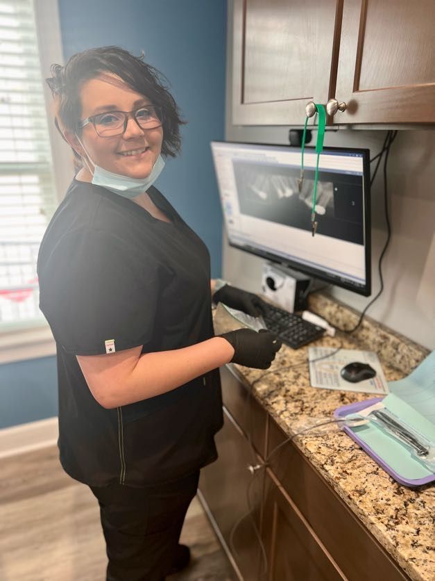 Dental assistant in black scrubs smiles, operating computer in office.