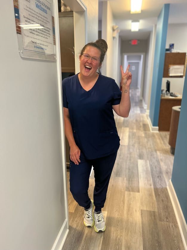 Woman in blue scrubs smiles and gives a peace sign in a hallway.