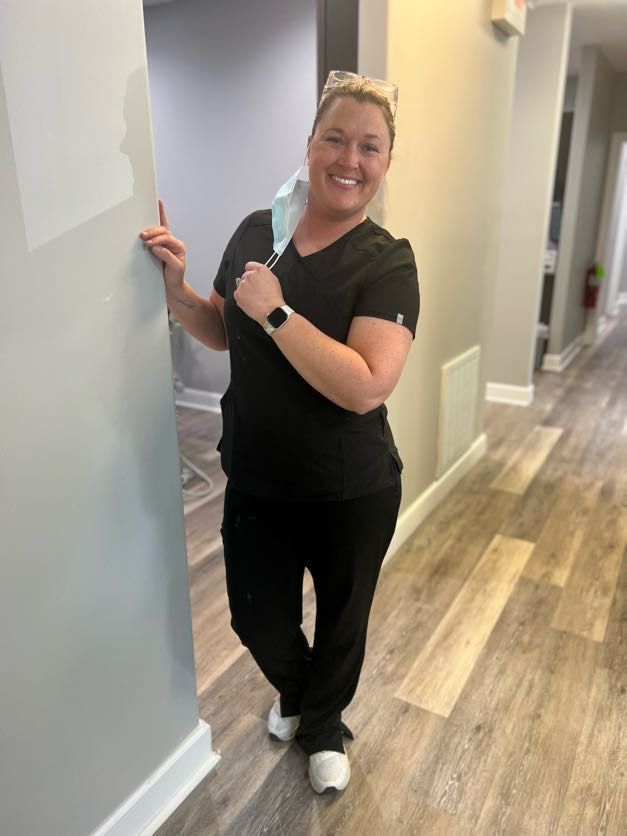 Woman in black scrubs smiles, holding a mask, standing in a hallway with wood-look flooring.