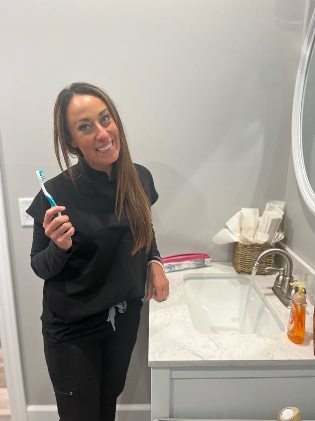 Woman in black scrubs holding a toothbrush, smiling by a bathroom sink and mirror.