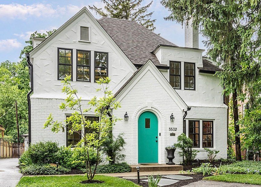 White brick house with black-framed windows and a turquoise door; lush green landscaping.