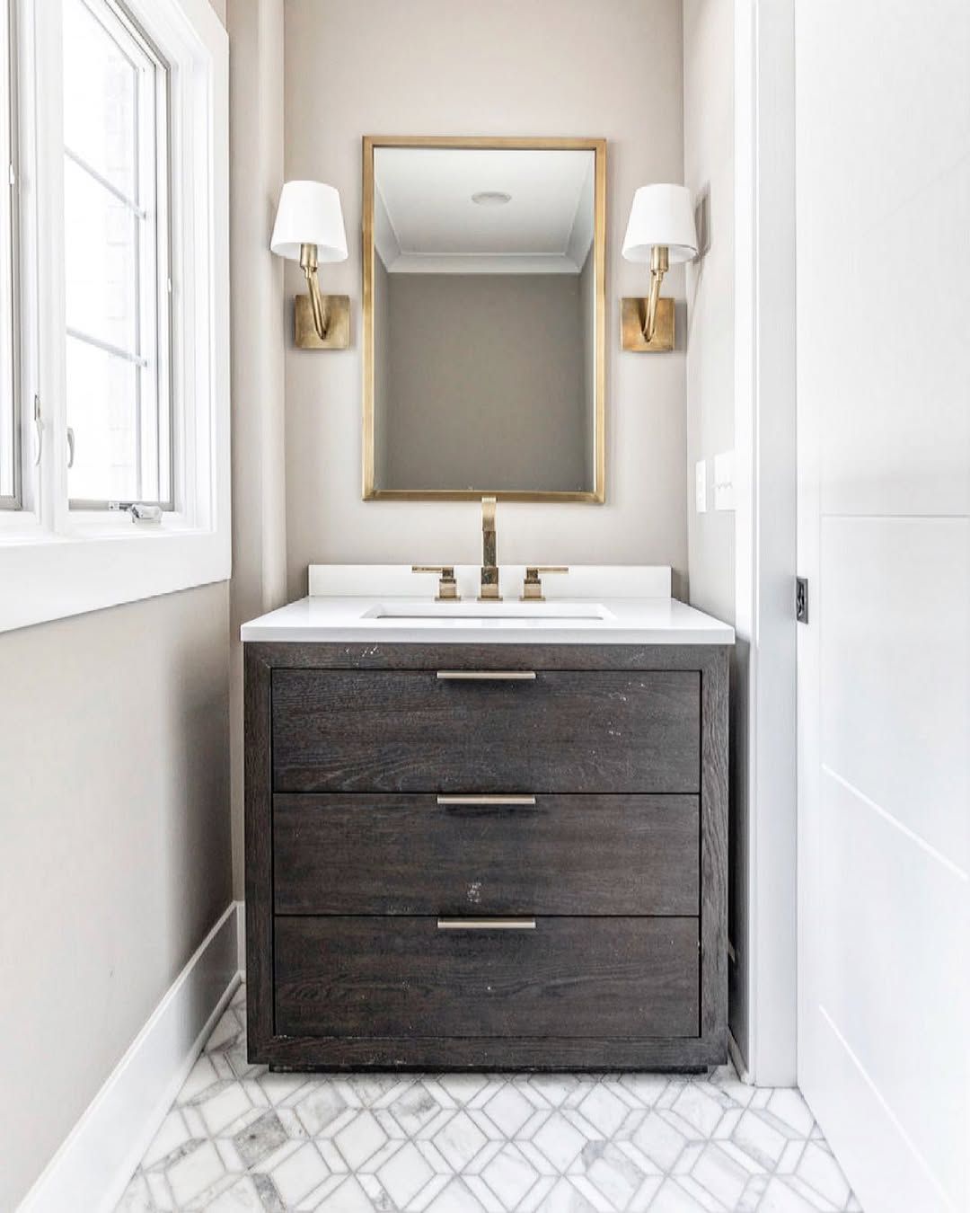 Small bathroom with dark wood vanity, gold mirror, sconces, and patterned floor.
