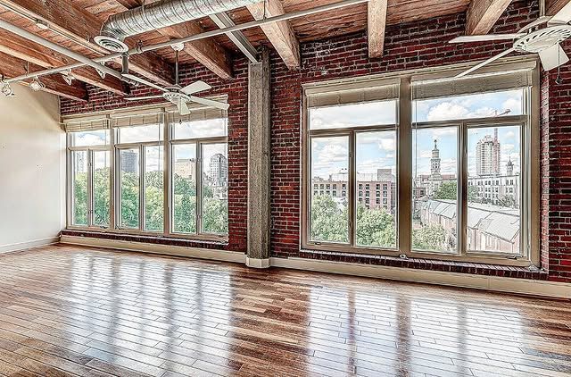 Empty loft space with brick walls, large windows, wooden beams, and city views.