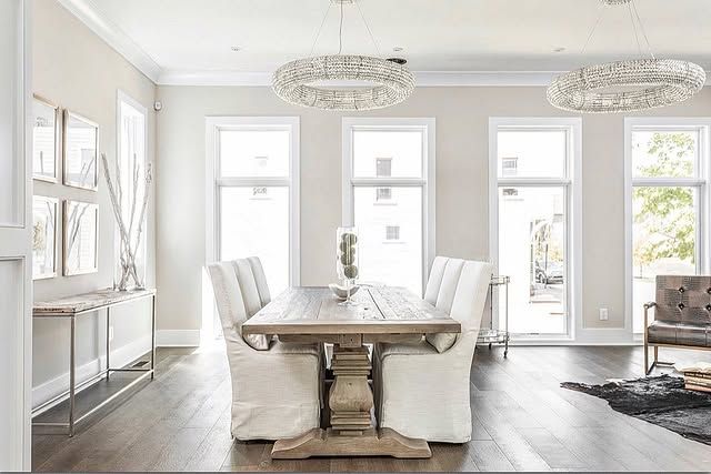 Dining room with a wooden table, white chairs, and bright windows. Two chandeliers hang above.