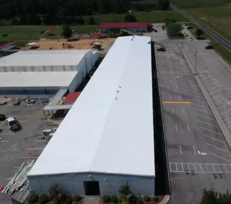 Aerial view of a long, white-roofed building with a parking lot, trees, and road on the right.