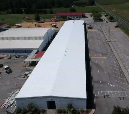 Aerial view of a long white building with a parking lot, trees, and a red-roofed structure in the background.