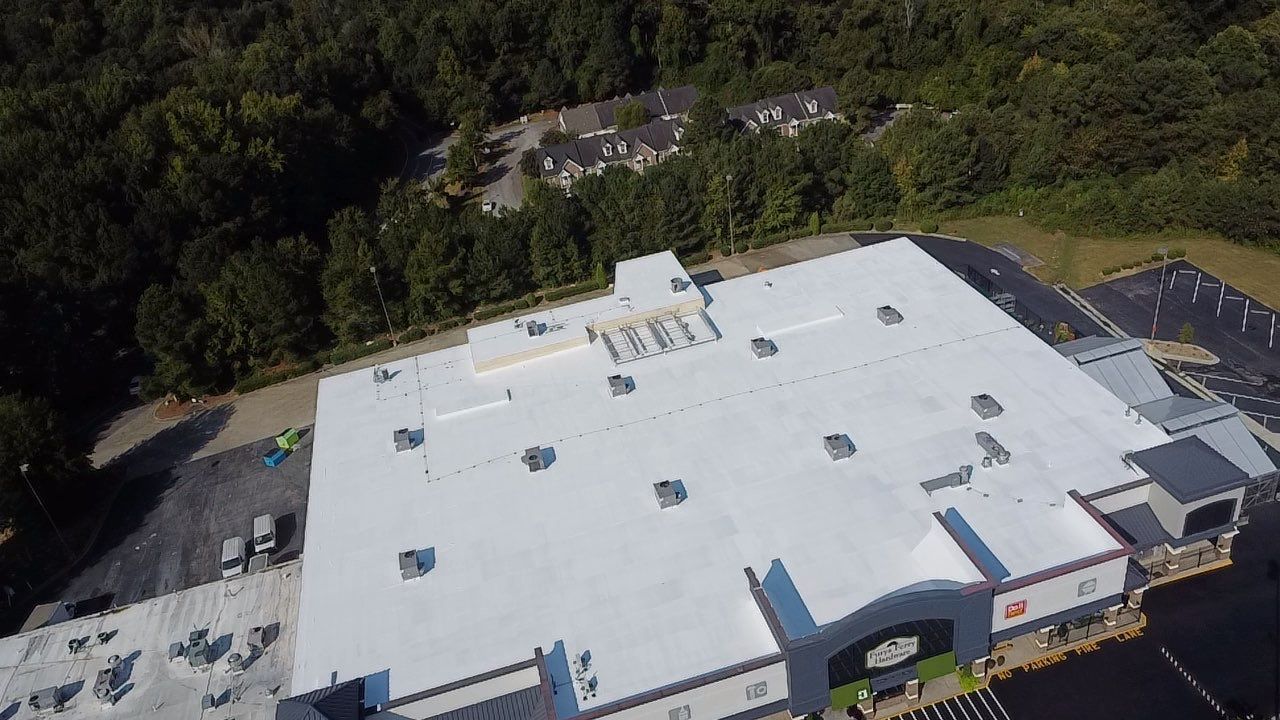 Aerial view of a large white commercial building roof with air conditioning units, surrounded by trees.