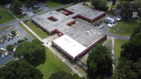 Aerial view of a brick school building with several wings and a white roof, surrounded by trees and parking lots.