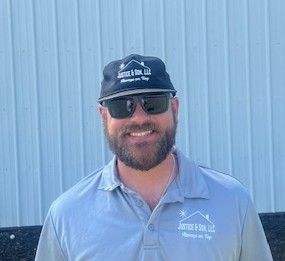 Man with beard, sunglasses, and hat, wearing a gray polo shirt, smiling outside.