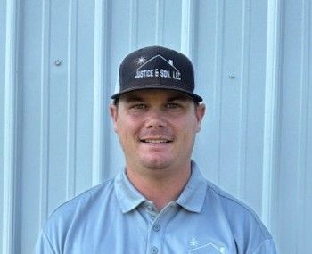 Man in a hat and polo shirt smiling in front of a light blue metal wall. The hat says 