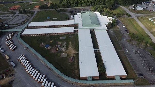 Aerial view of a large school building with white roofs, surrounded by parking lots, fields, and buses.