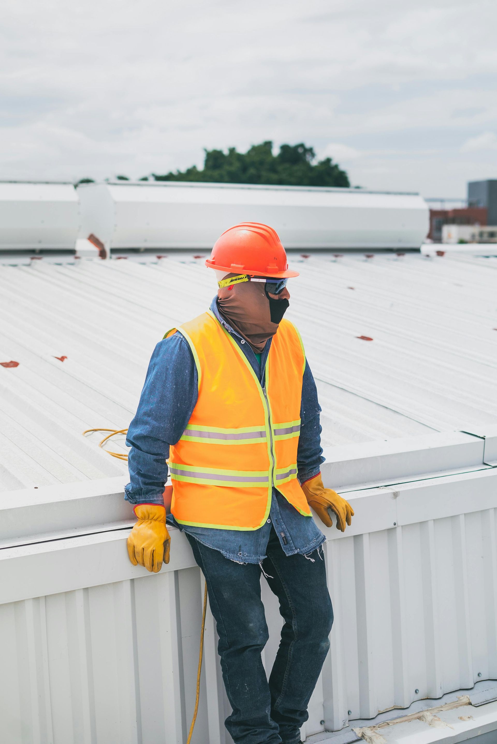 Construction worker on a rooftop, wearing orange safety gear and sunglasses, leaning, looking off to the side.