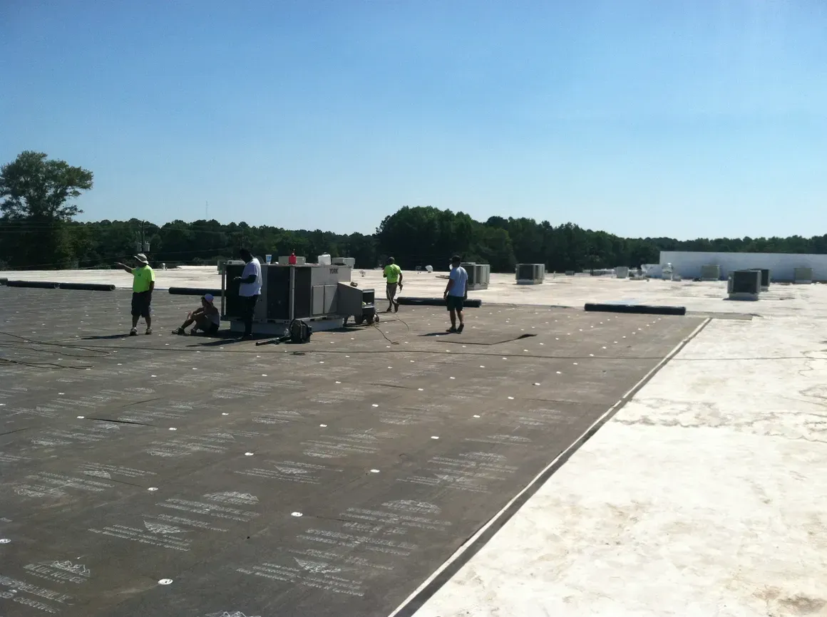 Workers on a flat roof, laying dark roofing material under a bright blue sky.