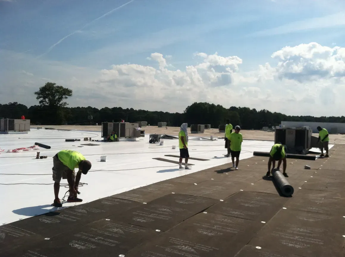 Roofers in neon vests working on a flat roof under a blue sky; some rolling out material.