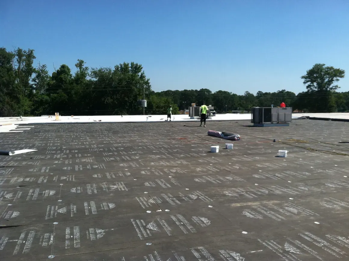 Workers on a black and white rooftop with equipment, trees, and a blue sky.