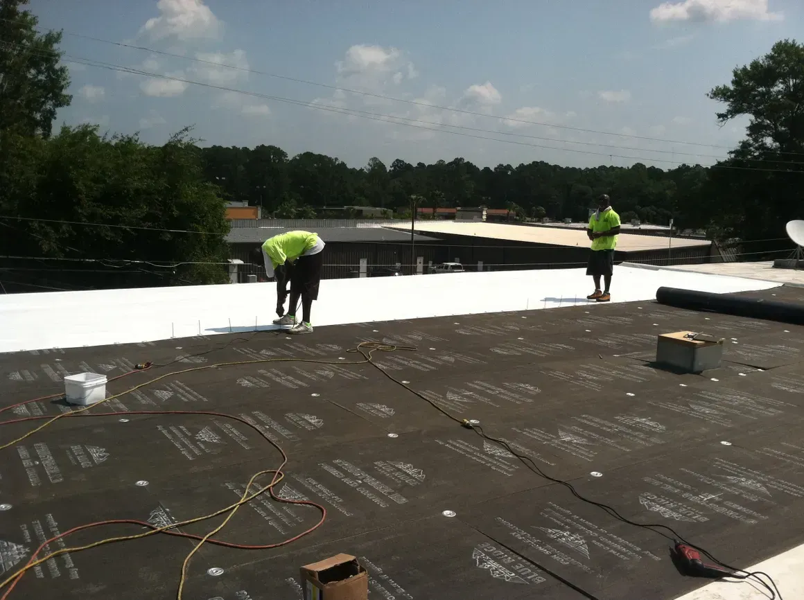 Two workers in neon vests install roofing material on a flat roof under a sunny sky.