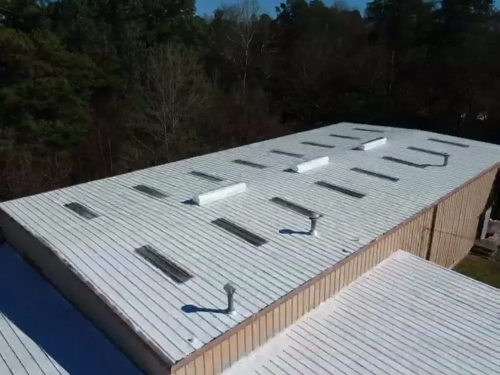 White metal roof of a building with rectangular skylights against a forest backdrop under a blue sky.