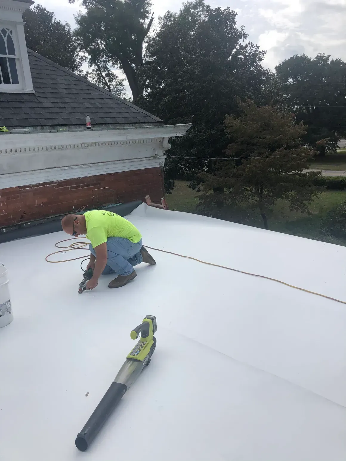 Man in green shirt working on a white flat roof, using a tool. A leaf blower is nearby.