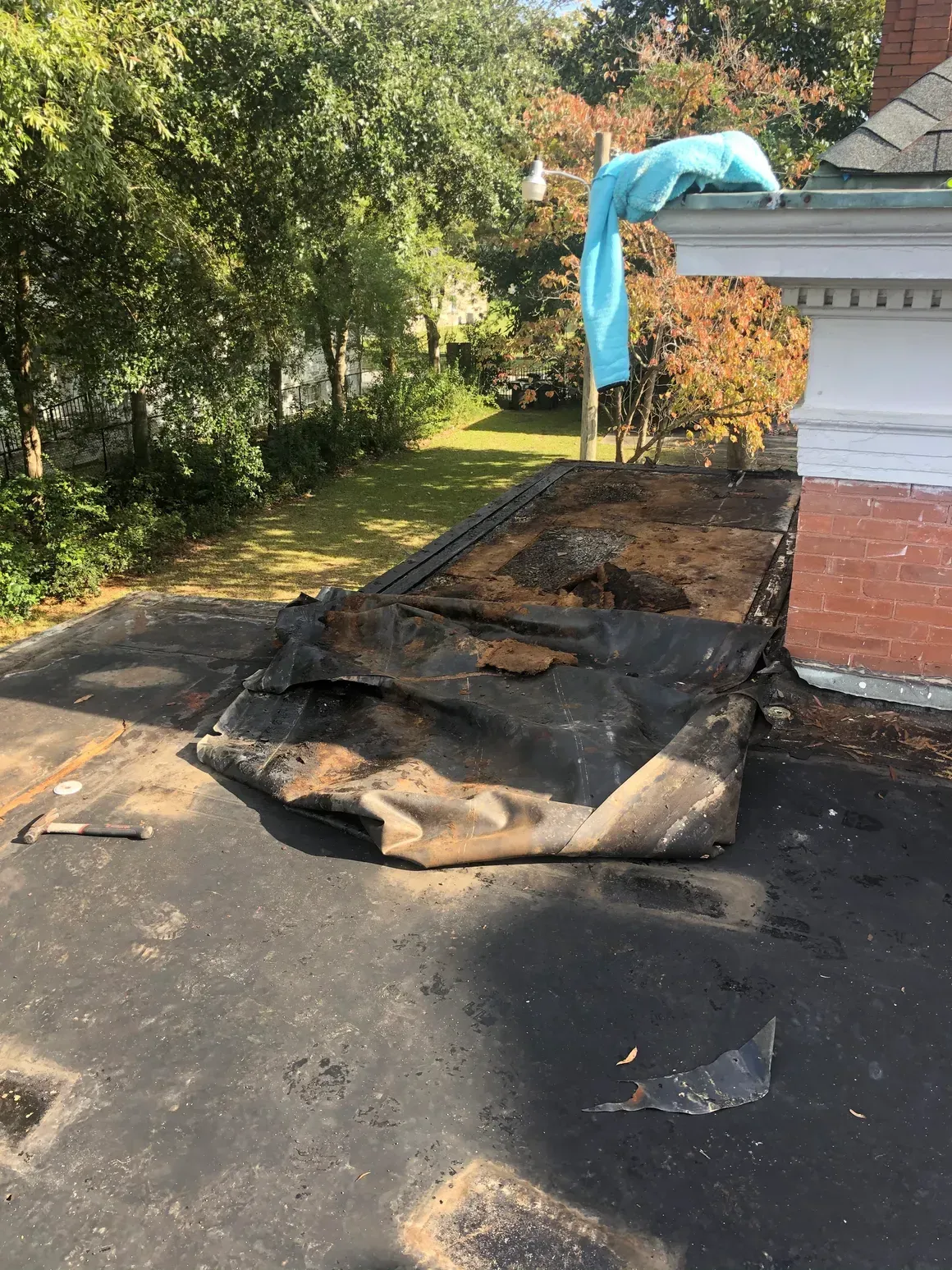 Damaged flat roof with torn black material and exposed underlayment next to brick chimney, outdoors.