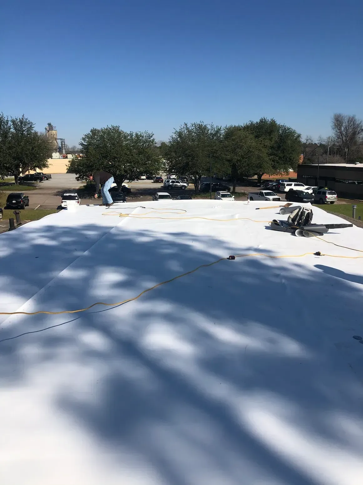 White roofing material being installed on a building with several vehicles and trees in the background.