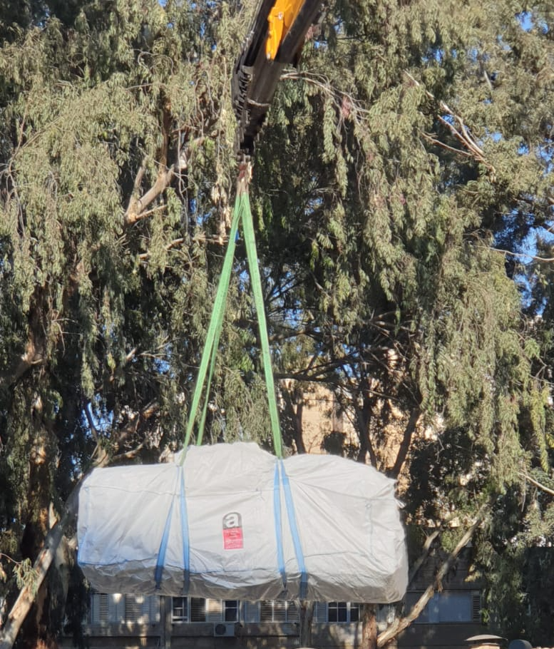 Crane lifting a large wrapped cylindrical load beneath a tree canopy.