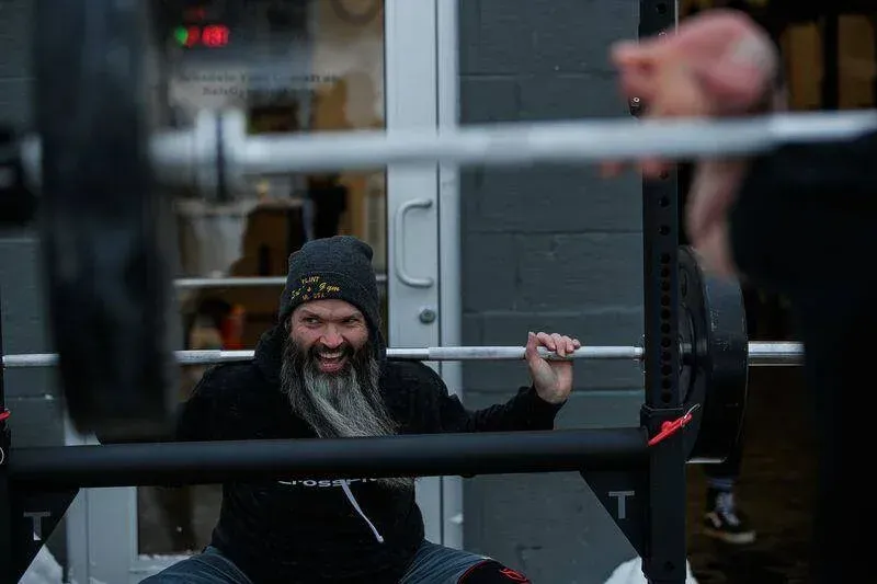 A man with a beard is squatting with a barbell in a gym.