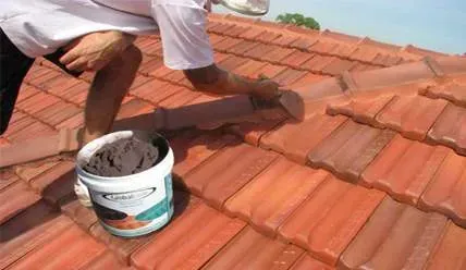 A man is kneeling on a roof next to a bucket of cement.