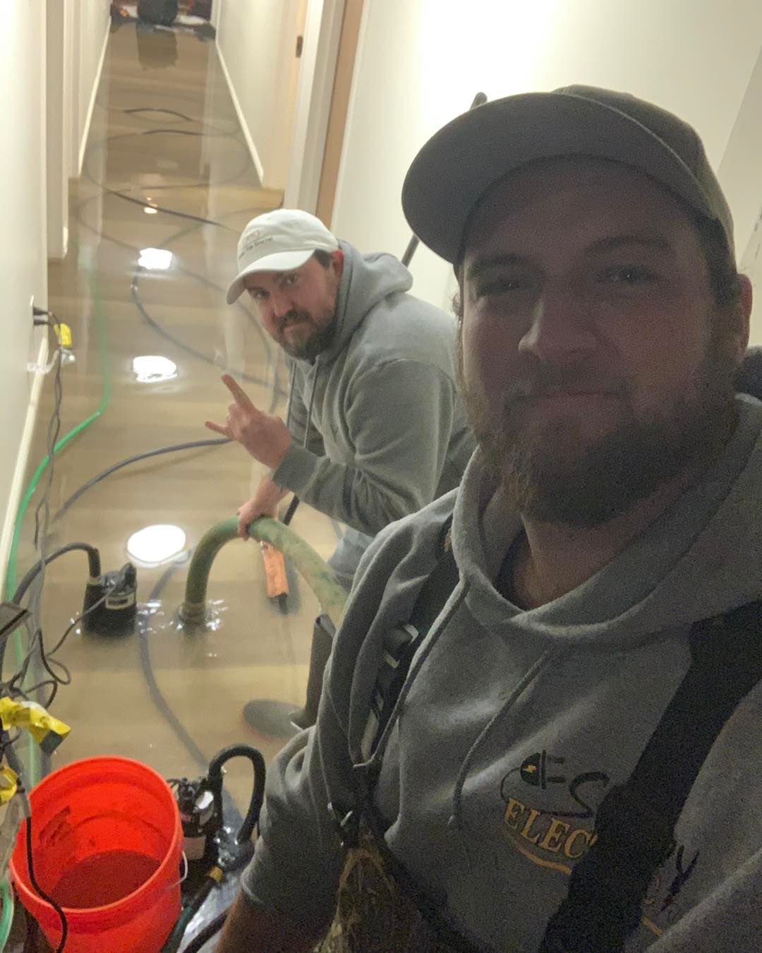 Two men in hoodies pumping water from a flooded hallway. One man takes a selfie.