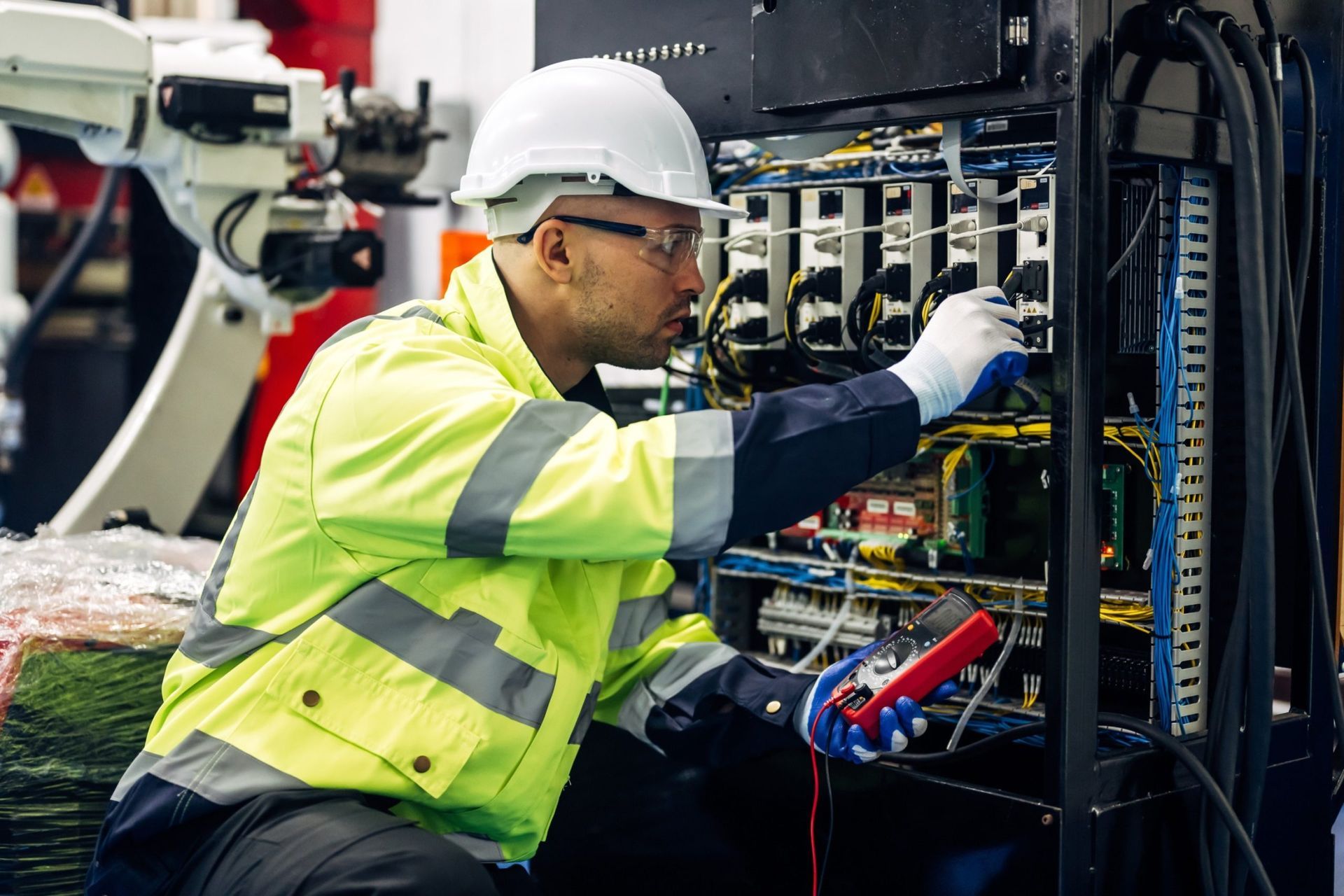 Man in safety gear inspecting machinery with a multimeter in a factory setting.