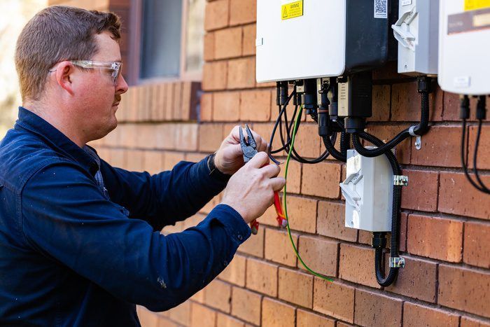 Electrician working on wiring near solar inverter boxes on a brick wall.