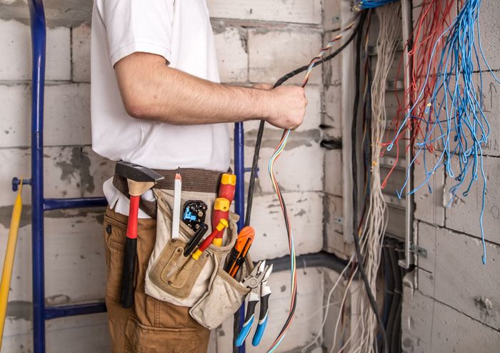 Electrician holding wires, working in unfinished room with tool belt and ladder.