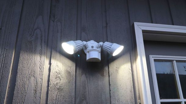 White outdoor security light with three adjustable heads on a gray wood-paneled wall, near a window.