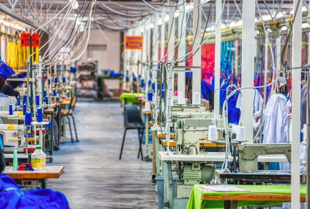 Sewing machines in a garment factory. Long rows, overhead wiring, fabric and spools of thread.