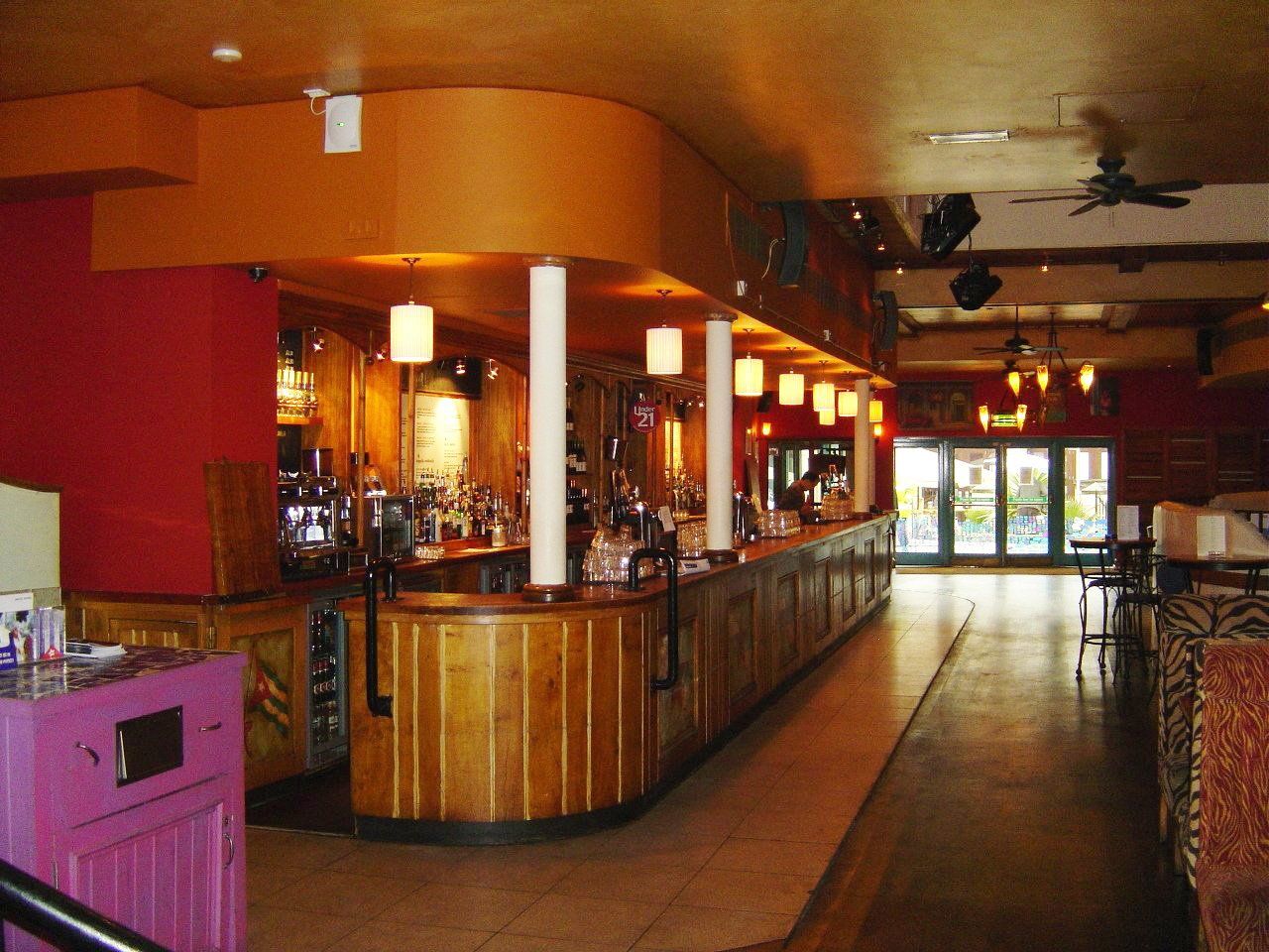 Interior of a bar with a long wooden counter, red and orange walls, and overhead lights.