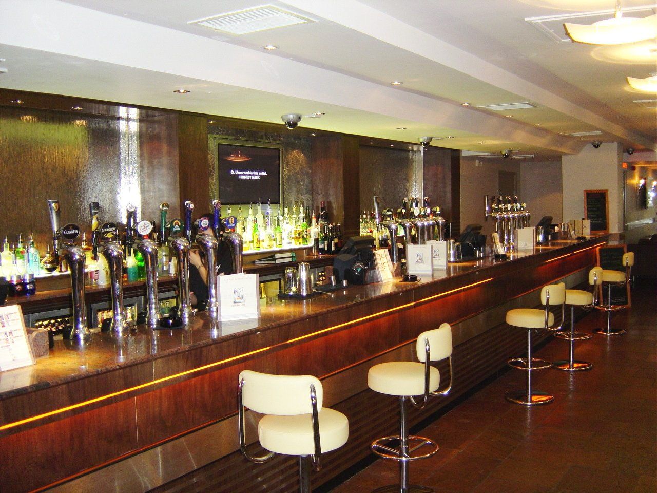 A bar interior with a long counter, bar stools, and taps against a wood and metal backdrop.