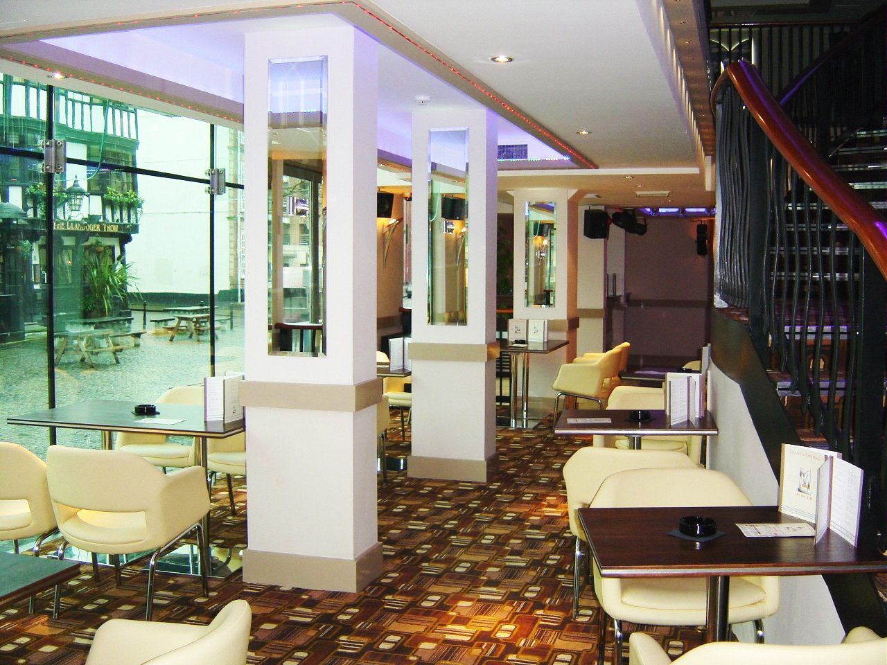 Interior view of a bar with cream chairs, brown tables, and a patterned carpet. Glass wall on the left.