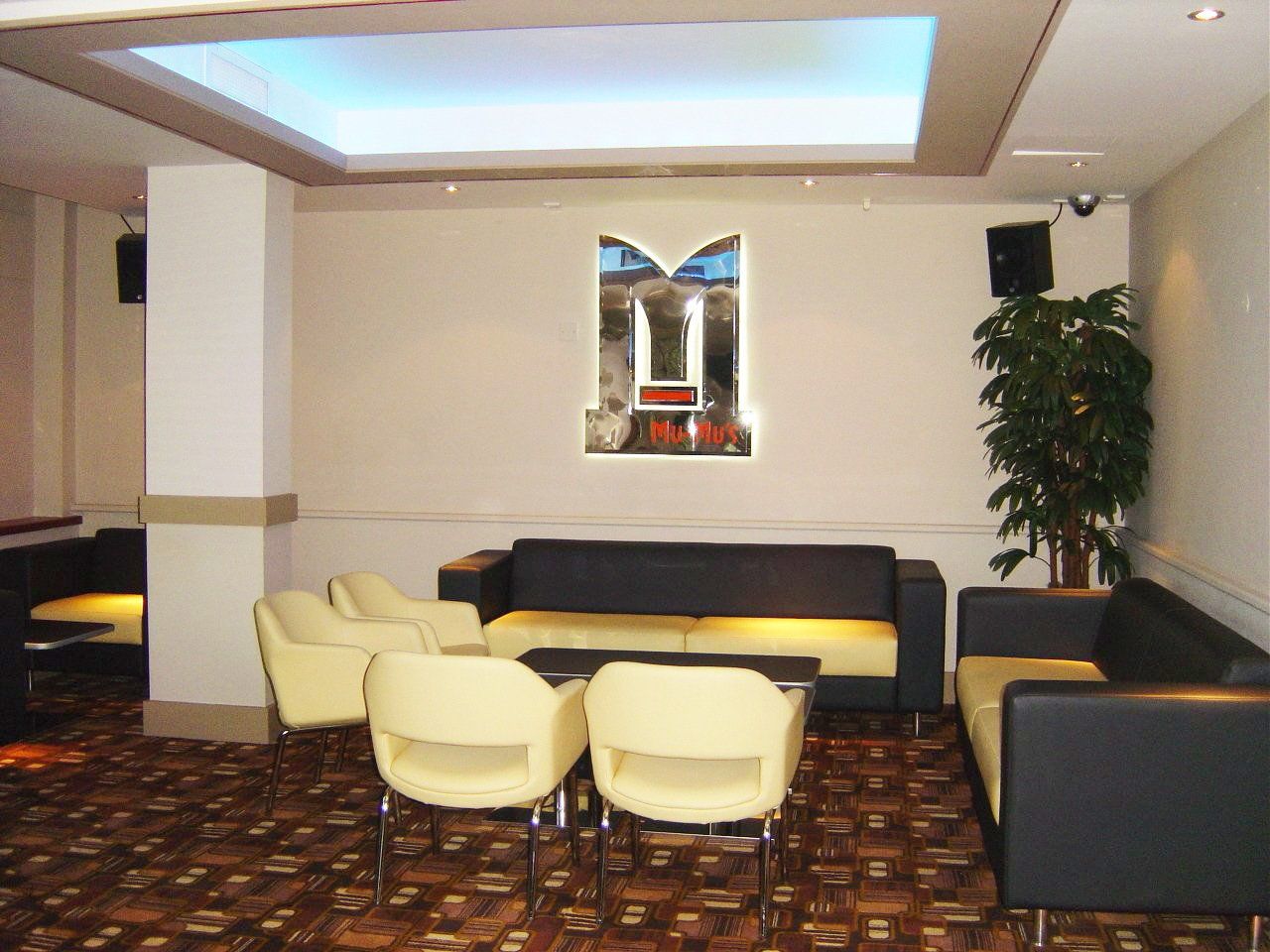 Lounge area with black and yellow sofas, cream chairs, patterned carpet, and a logo on the wall.