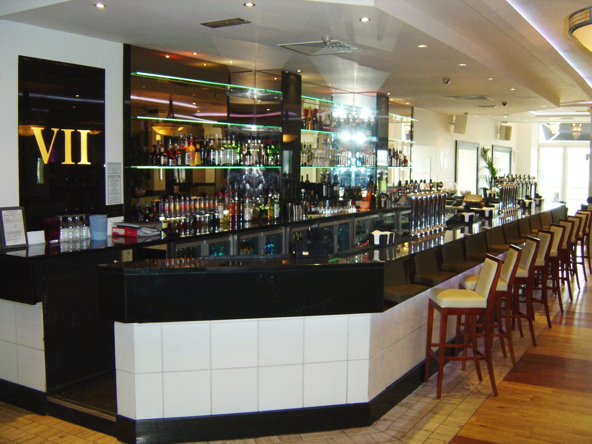 Bar with a long black countertop, stools, and well-stocked liquor display. The sign says