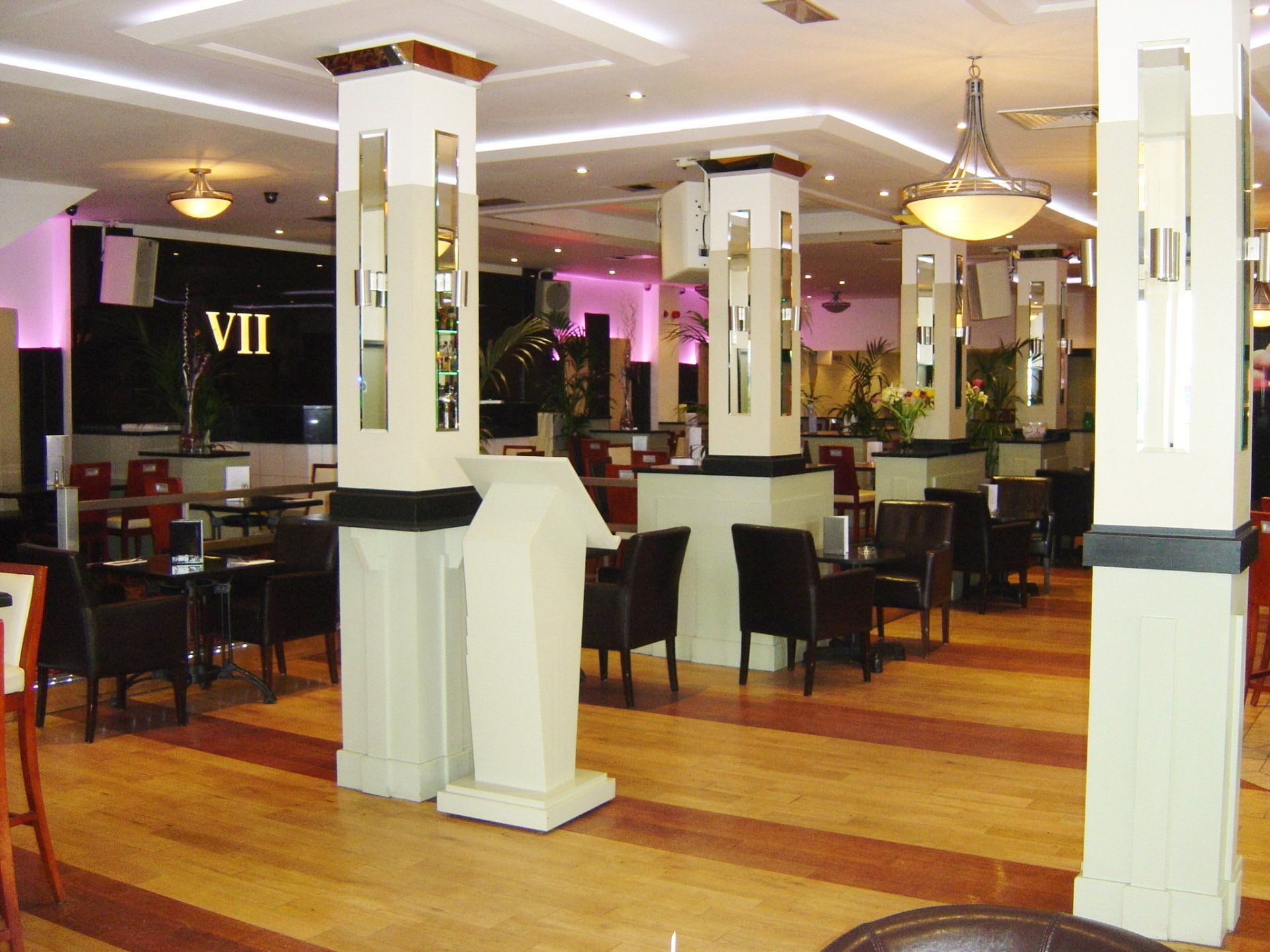 Interior of a restaurant with columns, tables, and chairs. Wooden floor with light fixtures, purple and yellow lighting.