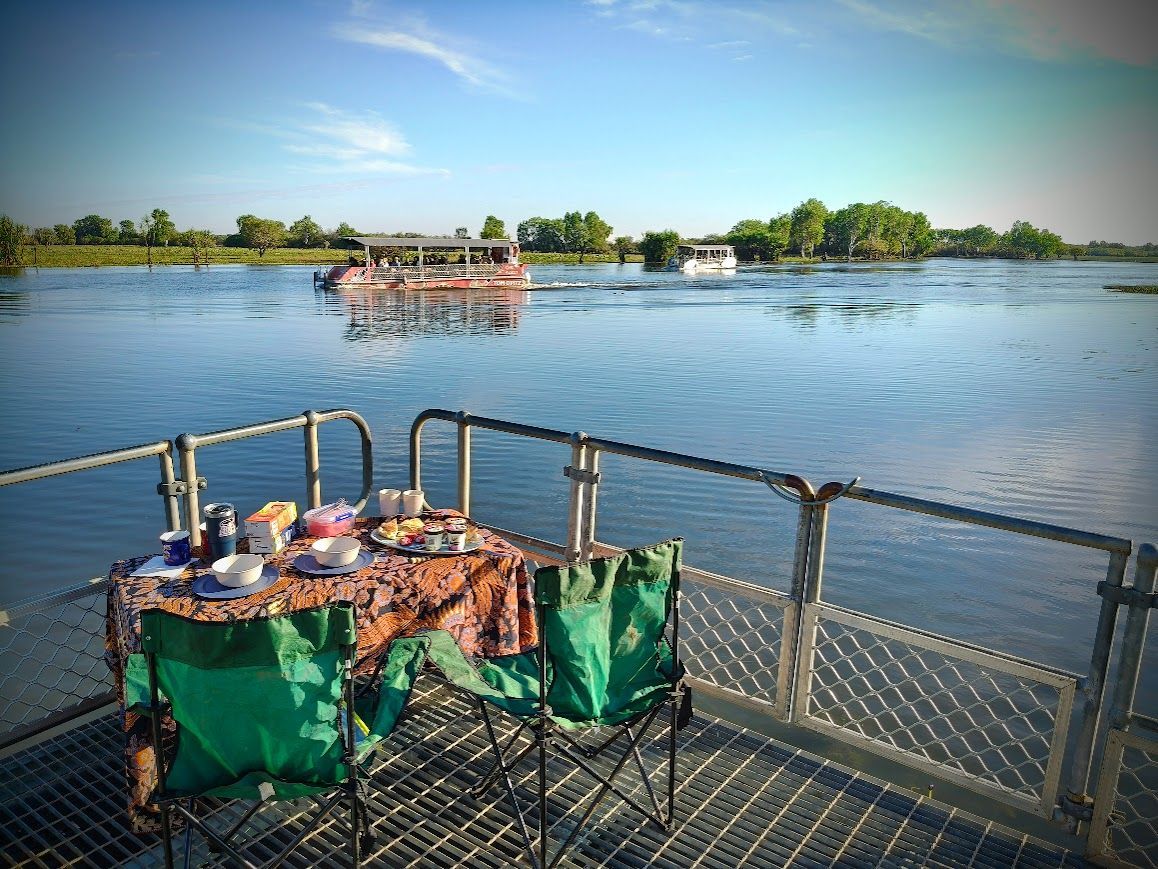 Picnic table set on a platform in a river, two boats in the distance. Blue water, green chairs, sunny day — NT Driver Guide - Escorted Tours & Transfers in Darwin City, NT