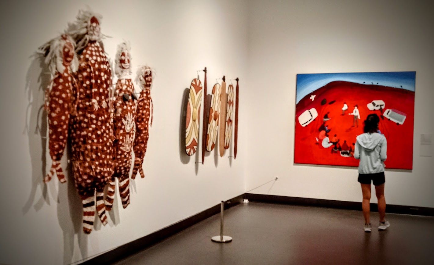 Art gallery interior with Indigenous Australian art on display; a person views a large red painting — NT Driver Guide - Escorted Tours & Transfers in Darwin City, NT