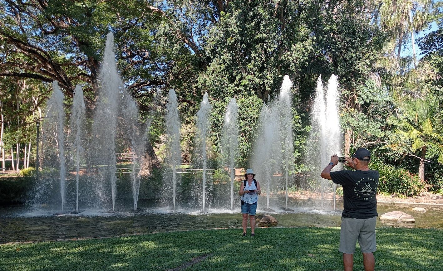 Fountain spraying water in park; two people near it, one taking a photo. Green grass and trees in background — NT Driver Guide - Escorted Tours & Transfers in Darwin City, NT
