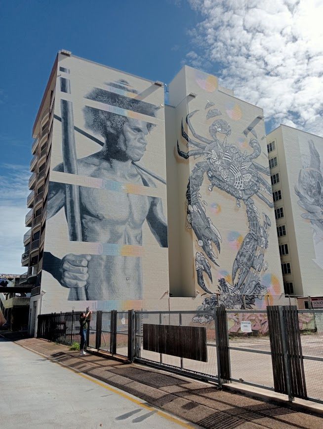 Large murals on buildings: Indigenous figure with spear; crab. Buildings against a blue sky — NT Driver Guide - Escorted Tours & Transfers in Darwin City, NT