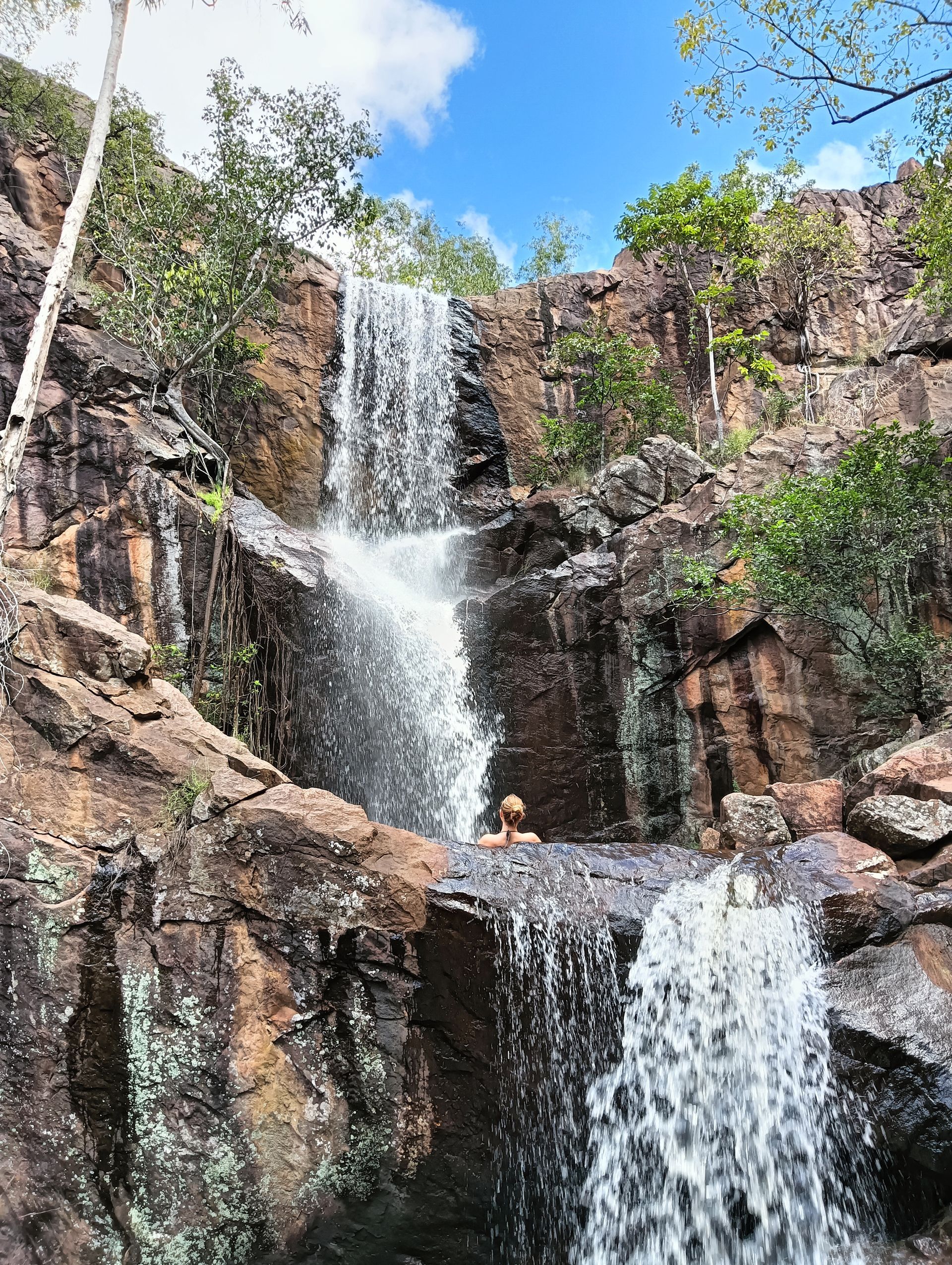 Waterfall cascading over rocky cliffs into a pool; blue sky with clouds in the background  — NT Driver Guide - Escorted Tours & Transfers in Darwin City, NT