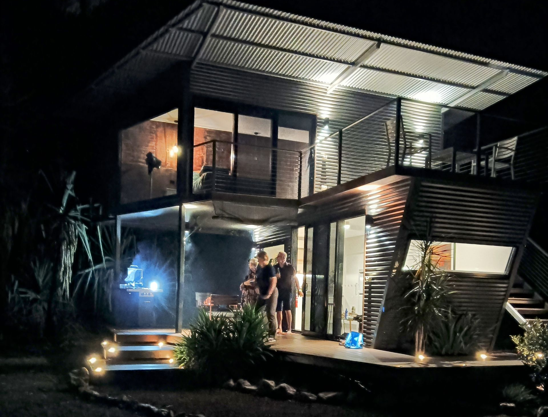 Modern two-story home illuminated at night with people gathered on the porch and a fire pit in the yard — NT Driver Guide - Escorted Tours & Transfers in Darwin City, NT