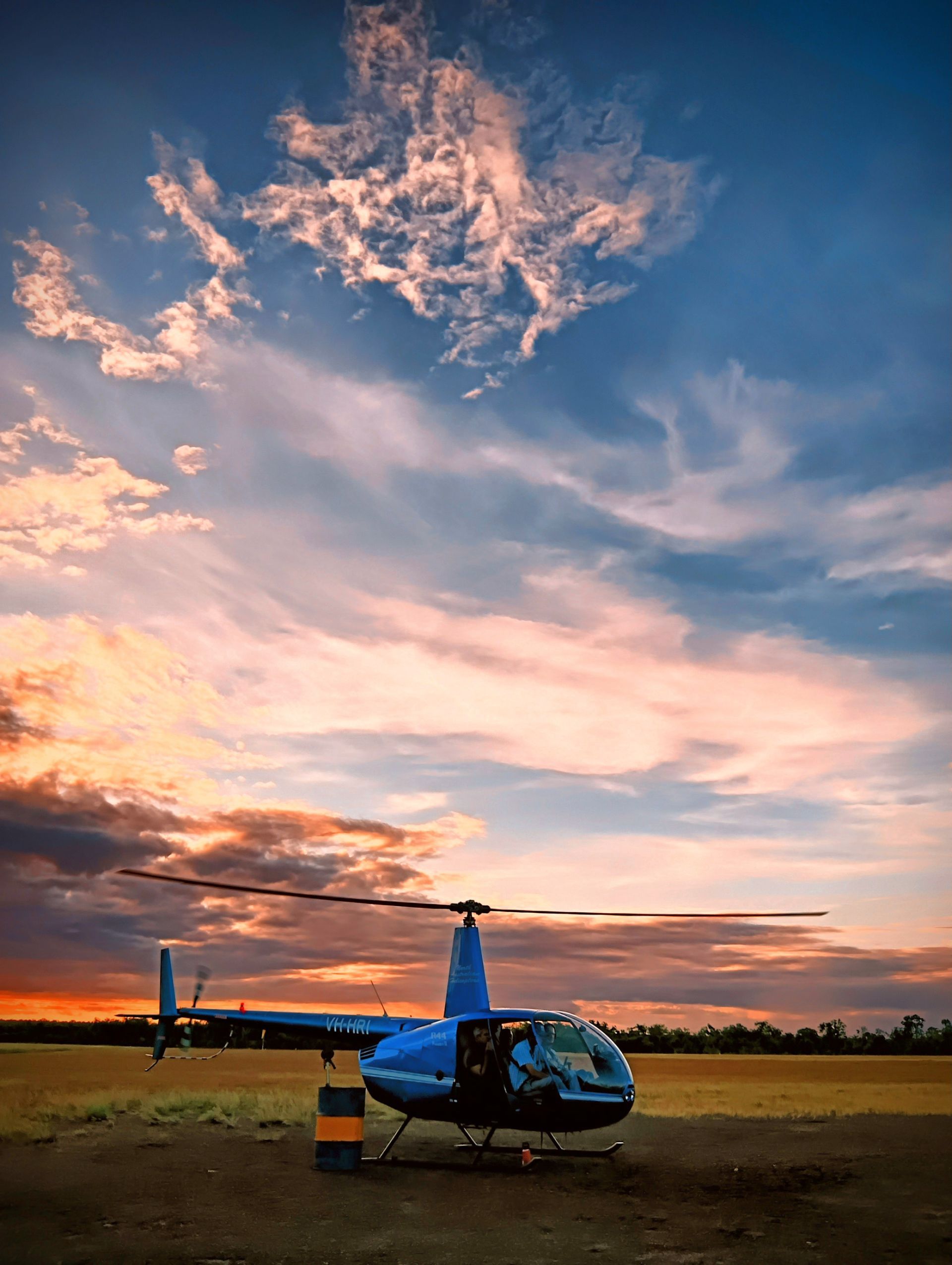 Blue helicopter parked on a grassy field at sunset, with colourful sky — NT Driver Guide - Escorted Tours & Transfers in Darwin City, NT